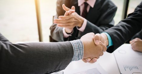 Two Business Men Shaking Hands In Front Of Man In Suit Clapping