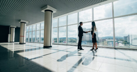 Businessman And Businesswoman Shaking Hands Together While Standing In Front Of Office Building Windows Overlooking The City